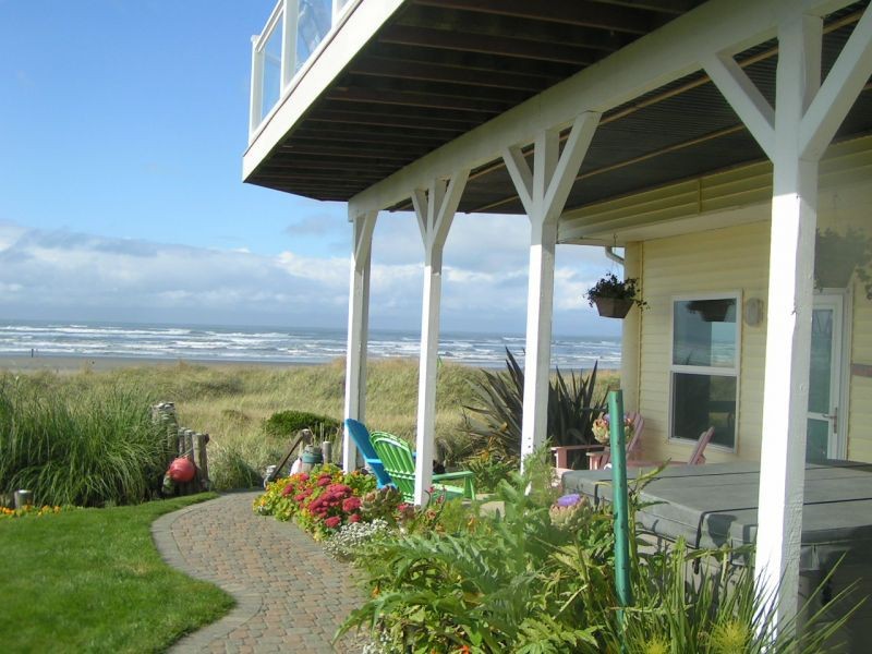 Yellow lower level beachfront home entrance with small landscaped yard, deck with chairs and brick walkway leading to a beach path. Ocean views and sandy beach.